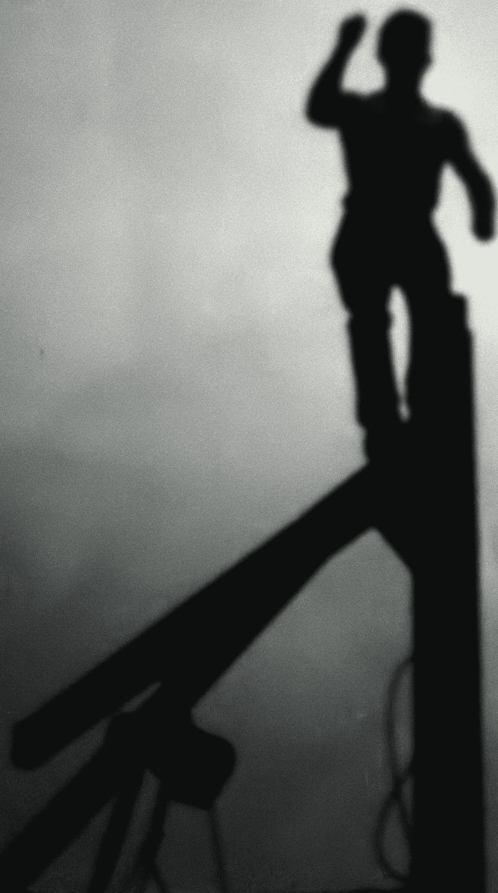 Black and white photograph: a young builder standing on top of a crane mast against a grey sky